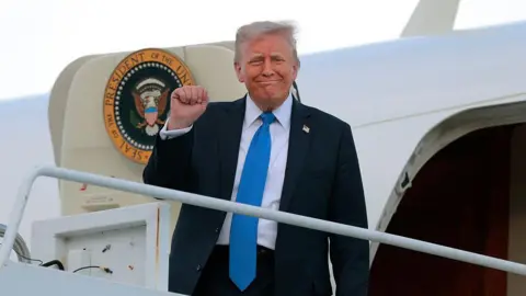 Getty Images Trump, wearing a dark suit and light blue tie, holds up a fist while boarding Air Force One to visit Alabama on Thursday