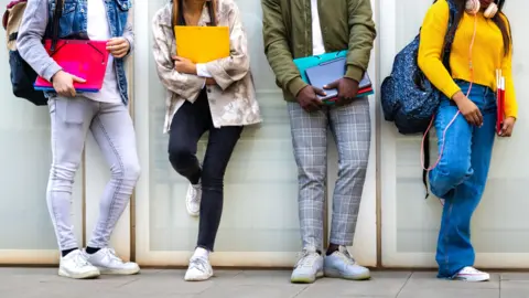 Getty Images Four students stand side by side. They hold folders and have rucksacks. Their faces are out of view.