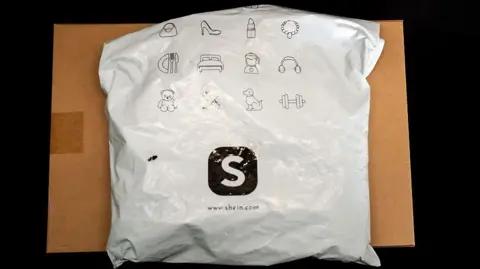 Getty Images A package from Shein, which is sitting on top of a cardboard box. The package is made of white plastic with black printing. the Shein S logo is prominent in the middle of the the parcel.
