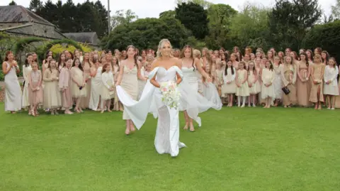 Mark Johnston A bride, with blonde hair, a white dress with an off the shoulder detail carrying a bouquet, smiles as two young bridesmaids help her with her train and veil as she walks towards the camera. Behind them is a large group of women, including young girls in the front row, wearing a mixture of champagne, cream, white and tan dresses. The crowd, including the bride and the two bridesmaid, are standing in the middle of a large garden, with tall trees and a leafy tunnel to the left.