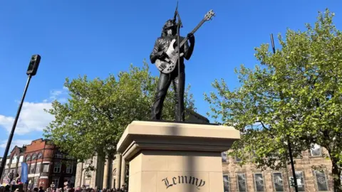 BBC A dark-coloured statue of Lemmy holding a bass guitar and signing upwards towards a microphone. It has been placed on a plinth, which reads "Lemmy", in the middle of a town square.