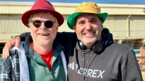 John Byford Meadows has his right arm around the artist as they pose for the photo. He has a pair of headphones round his neck and a hat to protect him from the strong sun. Byford wears glasses and a hat and both are grinning for the camera.