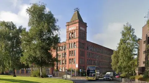 Google Redbrick four-storey mill with a five-storey tower that has a small mast at the top. Trees and vehicles can be seen on the roundabout in front.