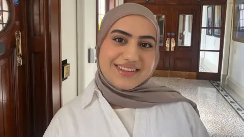 BBC A woman standing in a corridor, she is wearing a brown hijab with a white shirt.