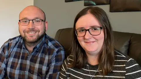BBC A balding man with a beard and glasses sitting on a sofa at home next to a woman with long brown hair and glasses