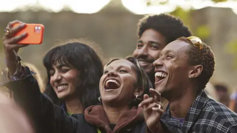 Getty Images A woman holds up a mobile phone in an orange case. She and her friends laugh and smiled for a selfie