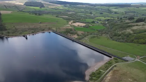BBC Drone image of Crai Reservoir. A large body of water can be seen in the foreground, surrounded by hills, fields and trees