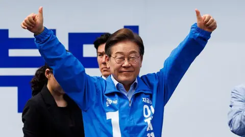 Getty Images Lee Jae–myung in a blue tracksuit jacket and glasses lifts both hands into the air with thumbs raised