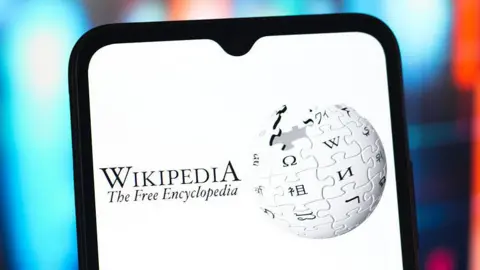 Getty Images A stock image showing a screenshot of Wikipedia's logo - a globe constructed out of jigsaw piece like elements bearing letters in different global alphabets - on a mobile phone. The text in the image reads "Wikipedia The Free Encyclopedia"