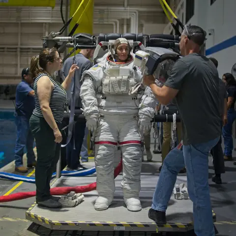 Kevin Church/BBC Astronaut Rosemary Coogan is standing on a platform next to a swimming pool at the Johnson Space Centre, Houston, Texas. She is wearing a white spacesuit - similar to the ones worn by the Apollo astronauts who landed on the moon. She's wearing a cap on her head that has a microphone attached to it. Two people are helping her. One woman in a striped vest and black trousers and a man in a black T-shirt and blue jeans who is holding the helmet that he is about to put over Rosemary's head. She is looking towards him. There are numerous other people in the background by the side of the pool.