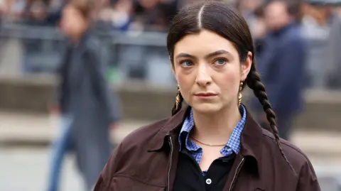 Getty Images Lorde pictured in March. The singer has her dark hair braided in two plaits either side of her head. She wears gold shell-shaped earrings and a brown jacket over a checked blue shirt. She's pictured outside, with people walking behind her in the distance.