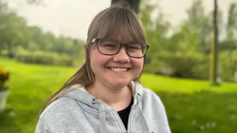 BBC Hannah Fullerton from Buglife has glasses, brown hair, tied back, and a fringe. She wears a grey hoody and is smiling widely.