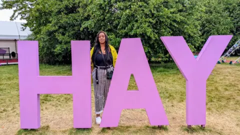 Jade Bradford Jade standing in the middle of big purple letters which spell out HAY. She is wearing a yellow jacket, black top and black and purple chequered trousers and there are trees behind her.