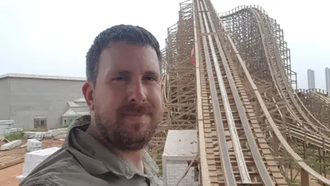 Marcus Gaines A man is stood at the bottom of a wooden rollercoaster, the track of which has a steep incline. He has a beard and is wearing a green top.