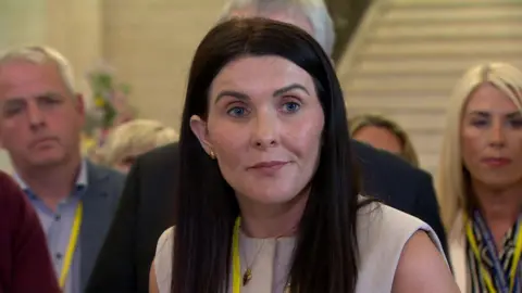 A woman is standing in front of a mic and talking to the media. There are other people standing around her. She has long brown hair and is wearing a yellow lanyard round her neck.