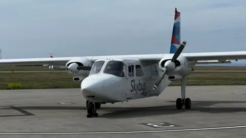 A small white Skybus plane parked on a runway at the airport. Grass and blue skies are visible in the background. 