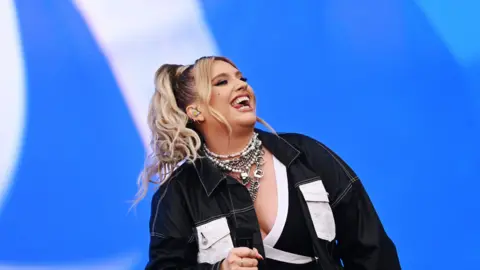 Ella Henderson performing at Radio 1's Big Weekend. She is smiling and has long blonde hair in a ponytail. She is wearing a black and white jean jacket, white and black top and lots of silver necklaces. 
