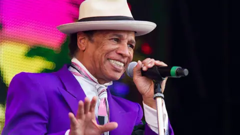 Redferns/Getty Images Kid Creole performing in 2015.  He is wearing a purple suit with a pink tie and white hat. He is holding onto the microphone while singing into it.