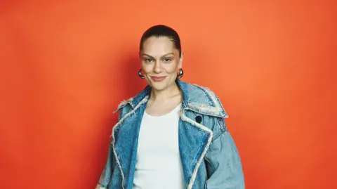 Jonathan Birch/BBC Jessie J is standing against an orange background and smiling at the camera. Her black hair is tied back and she is wearing silver hoop earrings. She is also wearing a white top and jean jacket. 