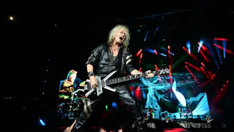 Paras Griffin/Getty Images Joe Elliott  is playing guitar on stage while singing. He is dressed in all black leather and has medium length white hair. The stage is dark with some bright blue and red lights shining across it. 