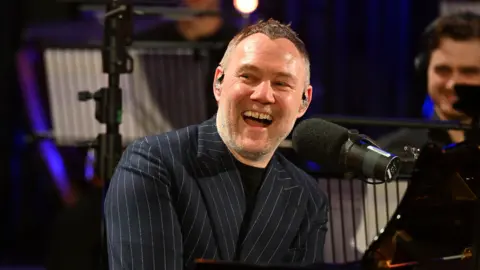 Mark Allan/BBC David Gray is sitting at a black piano and laughing into a microphone. He has grey stubble and grey hair. He is wearing a navy blue suit jacket with pin stripes. 