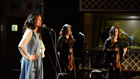 Mark Allan/BBC Sophie Ellis Bextor is singing into a microphone at a recording studio. She has her hands on her hips and eyes closed. She has long brown hair and is wearing headphones and blue dress with white sleeves. Next to her are two female backing singers. 
