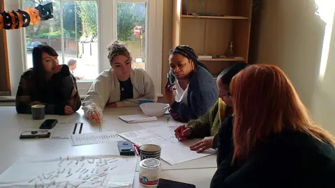 Link to Change A group of people in a room, looking at paper. There are five women in all, one is writing, and they are all concentrating. 