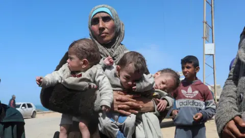 Reuters A Palestinian woman carries three babies in central Gaza after fleeing al-Shifa hospital in Gaza City, on 21 March 2024