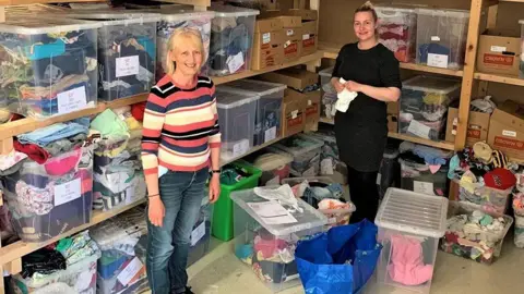 Leeds Baby Bank Two women stand next to racks and boxes of baby clothes. 