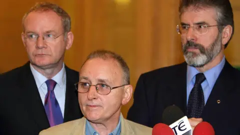 PA Media Martin McGuinness, Denis Donaldson and Gerry Adams are all looking to their right while speaking at a press conference. All three are wearing glasses. Mr McGuinness and Mr Adams are both wearing dark suits, shirts and ties. Mr Donaldson is wearing a tanned coloured jacket and a denim shirt. There are three microphones in front of them.