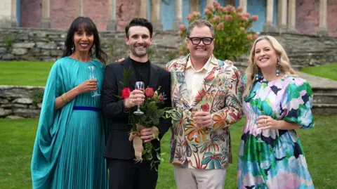 BBC/DSP/Georgina Vincent John Cooper wears a black suit and stands holding flowers and a glass of champagne next to Alan Carr. Elegantly-dressed women stand on either side of them, also each holding glasses of champagne. 