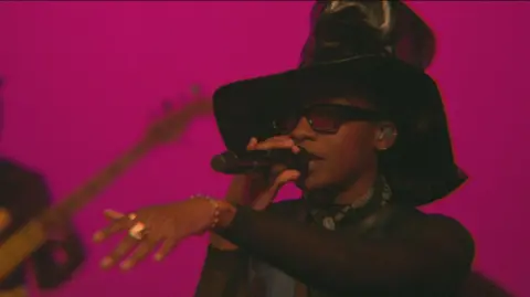 Little Simz performing at the BBC's Maida Vale studios. She wears an oversized black leather top hat and neck scarf and holds out her left hand while rapping. She's pictured against a pink backdrop and a bassist can be seen in the background