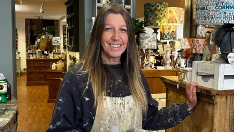 A woman with chest length brown hair. She is wearing an apron that is covered in paint and a blue sweater that has paint of the sleeves. Around here is itends from her shop. To the right a wooden chest of drawers with a lamp and to the right a grey chest of drawers with notebooks on. 