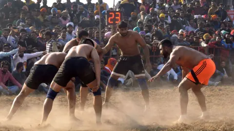 Getty Images Baba Suckhchain Dass Kabaddi Shahkot (R) attempts to tag an Dashmesh Kabaddi Club Nakodar opponent during the first Kabaddi Cup Tournament in Amritsar on February 5, 2016. Five men wearing shorts and some wearing knee grips lean in to a scrum formation at a dusty sports ground watched by a big audience. 