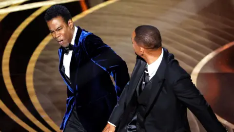 Getty Images Will Smith slaps Chris Rock onstage during the show at the 94th Academy Awards at the Dolby Theatre at Ovation Hollywood on Sunday, March 27, 2022. Chris Rock, wearing a blue velvet suit and bow tie, grimaces after being struck. Will Smith, pictured from the back, wears a black tux, his right arm in front of him after striking the comedian on stage. 