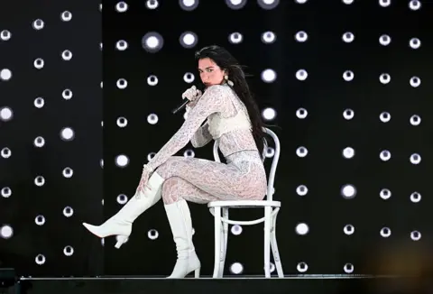 Getty Images Dua Lipa dressed in a white lace bodysuit performs a song while sitting on a chair