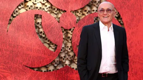 Getty Images Boyle is wearing a white shirt, black suit jacket and glasses. He is standing in front of a red background with the film's warning sign logo, which features mocked-up human skulls.