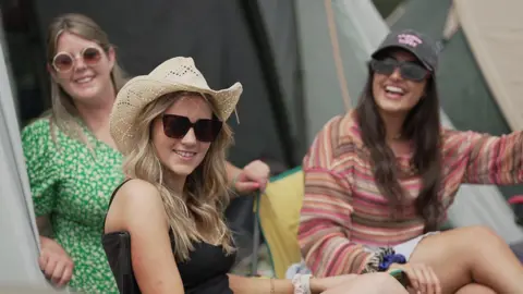 A girl wearing a straw hat and sunglasses smiles at the camera alongside two women 