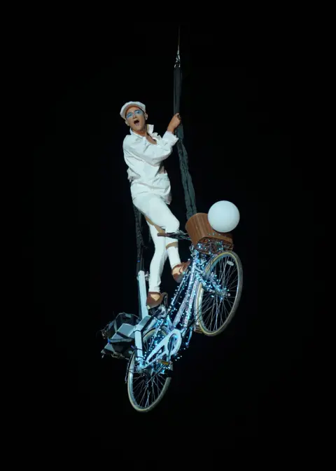 Yui Mok/PA A man in white clothing stands on a lit-up bike, which is suspended in the air. 