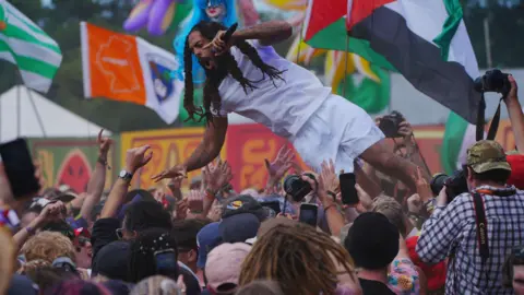 PA Media A man is about to land in a crowd after jumping from the stage. He is wearing a white shirt and shorts and has long, dark hair. 