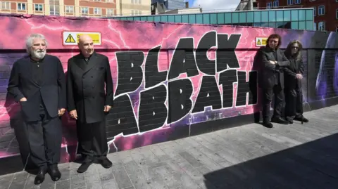 Darren Quinton/Touchstone Visuals The four original members of Black Sabbath four older men, all who are dressed in black, standing in front of a street art mural that reads "Black Sabbath" in large black bubble writing. The background of the mural is pink and purple and features white lightning strikes. 