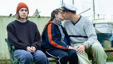 BBC/Various Artists Limited/Vishal Sharma From left to right: Sitting on a bench Josie (Sadler) is wearing blue trousers and a blue jumper with an orange beanie. She is sitting next to Billie (Davidson) wearing a blue tracksuit with a striped top and is kissing Nicky (Sam Buchanan) who is wearing green trousers a grey jumper and a sea captain's hat. 