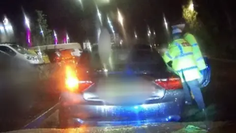 Police Scotland The dashcam footage shows a night-time scene. Two police officers, wearing high visibility jackets, are at the open driver's door of a blue car.