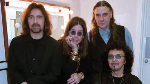 BBC Butler, Osbourne, Ward and Iommi, dressed in black, sitting around a lit dressing room mirror. Their expressions are solemn as they look at the camera.