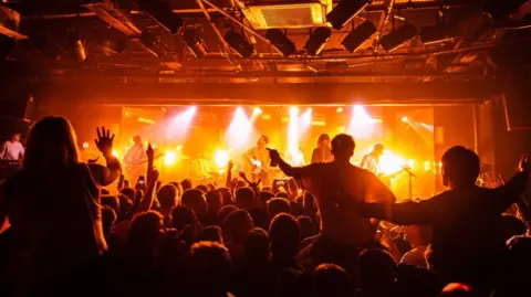 Ewan Ogden A general view from the back of the Leadmill music venue. A man, Miles kane, is on stage in the distance and a crowd with people dancing and sitting on each others shoulders is lit under orange lights