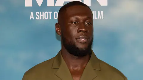Louis Browne Stormzy at the Big Man premiere. He has a short beard and shaved head and wears a mossy green shirt. He's pictured in front of a board printed to resemble a blue sky.