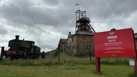 BBC A picture of the Big Pit Museum with the red welcome sign to the right and a black steam train on the left. The day is cloudy. 
