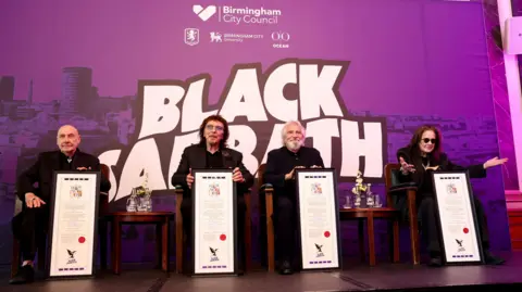 Birmingham City Council From left to right, Bill Ward, Tony Iommi, Geezer Butler and Ozzy Osbourne receive their scrolls. There is a council branded Black Sabbath board behind them in purple. They are sat in front of it on chairs. All are wearing black. Ozzy has his arms outstretched. 