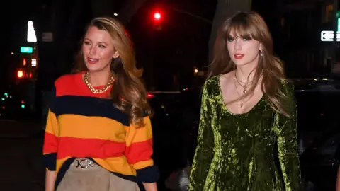 Getty Images Blake Lively, wearing an orange, red and black stripy jumper and a tan suede skirt, walking alongside Taylor Swift, who is in a green crushed velvet dress