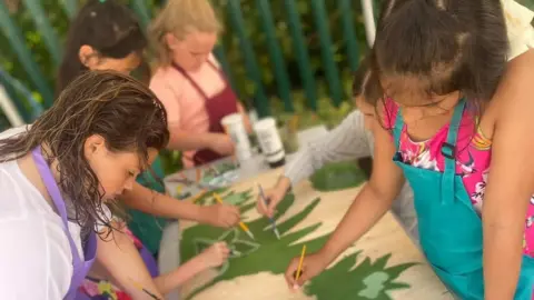 YACAD A group of young girls painting a large piece of artwork which is laid out on a table in front of them. It's a shared activity with them all working on the same piece. They hold small paintbrushes and wear colourful aprons.